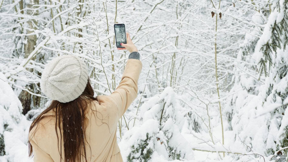 Woman in winter coat and hat taking a photo with her phone in a snow-covered forest.