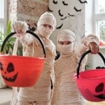 Three children dressed as mummies holding Halloween-themed buckets in a cozy, decorated room.