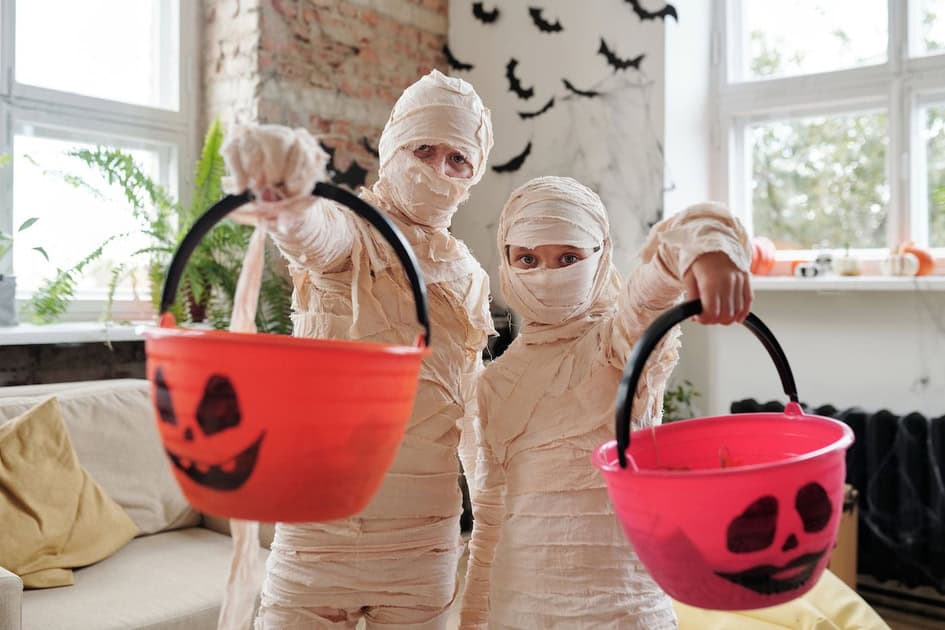 Three children dressed as mummies holding Halloween-themed buckets in a cozy, decorated room.