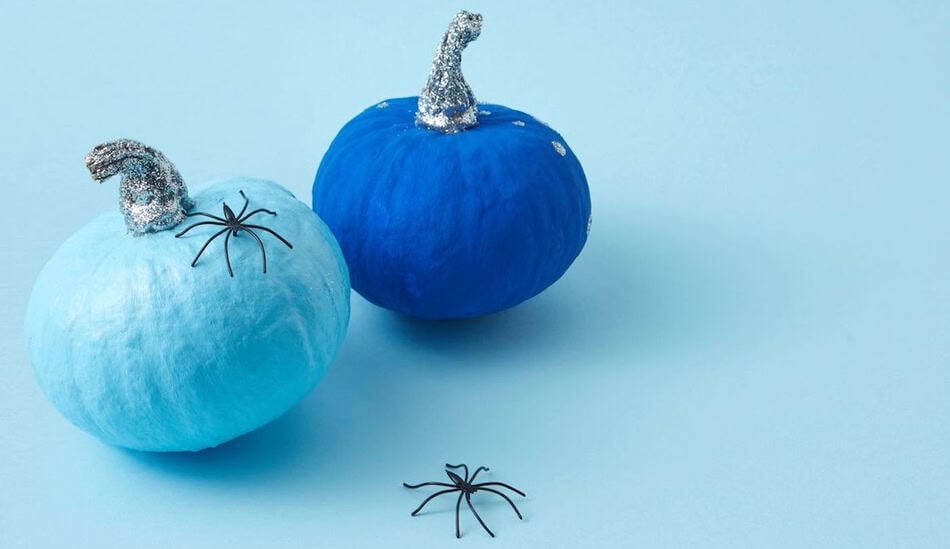 Teal pumpkin with glittery witch hat and plastic spiders on light blue background.