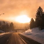 Bright sun setting over a snowy landscape with trees and a highway.