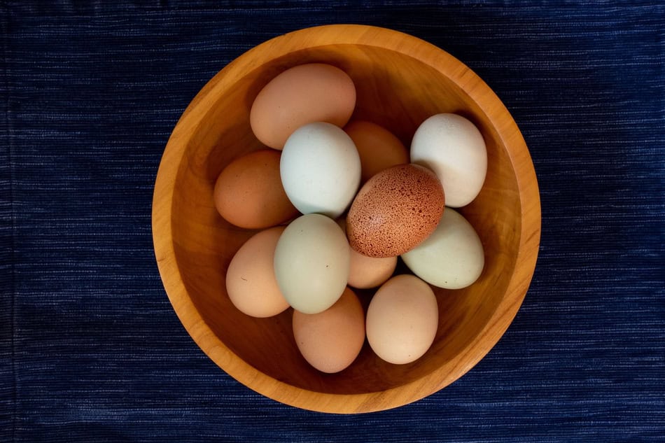 Assorted fresh eggs in a wooden bowl on dark fabric background.