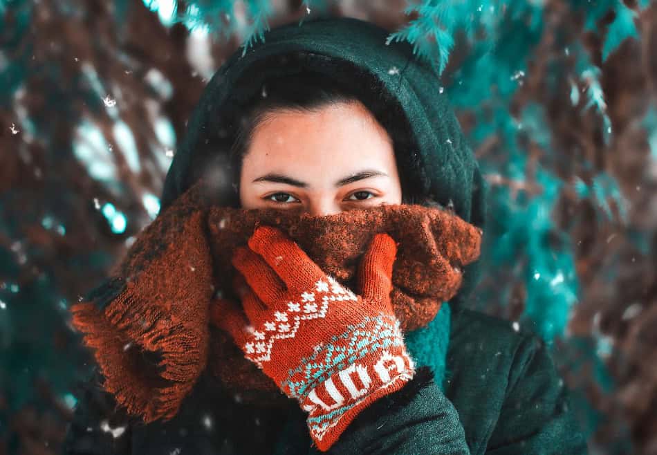Woman in winter clothing covering her face with gloves in snowy outdoor setting.