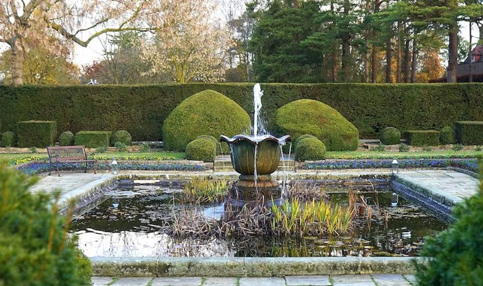 Elegant garden fountain surrounded by trimmed bushes and trees in a landscaped park setting.