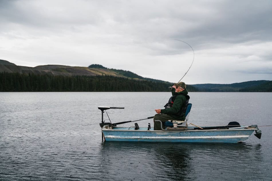 Man fishing in a small boat on a calm lake with mountains in the background.