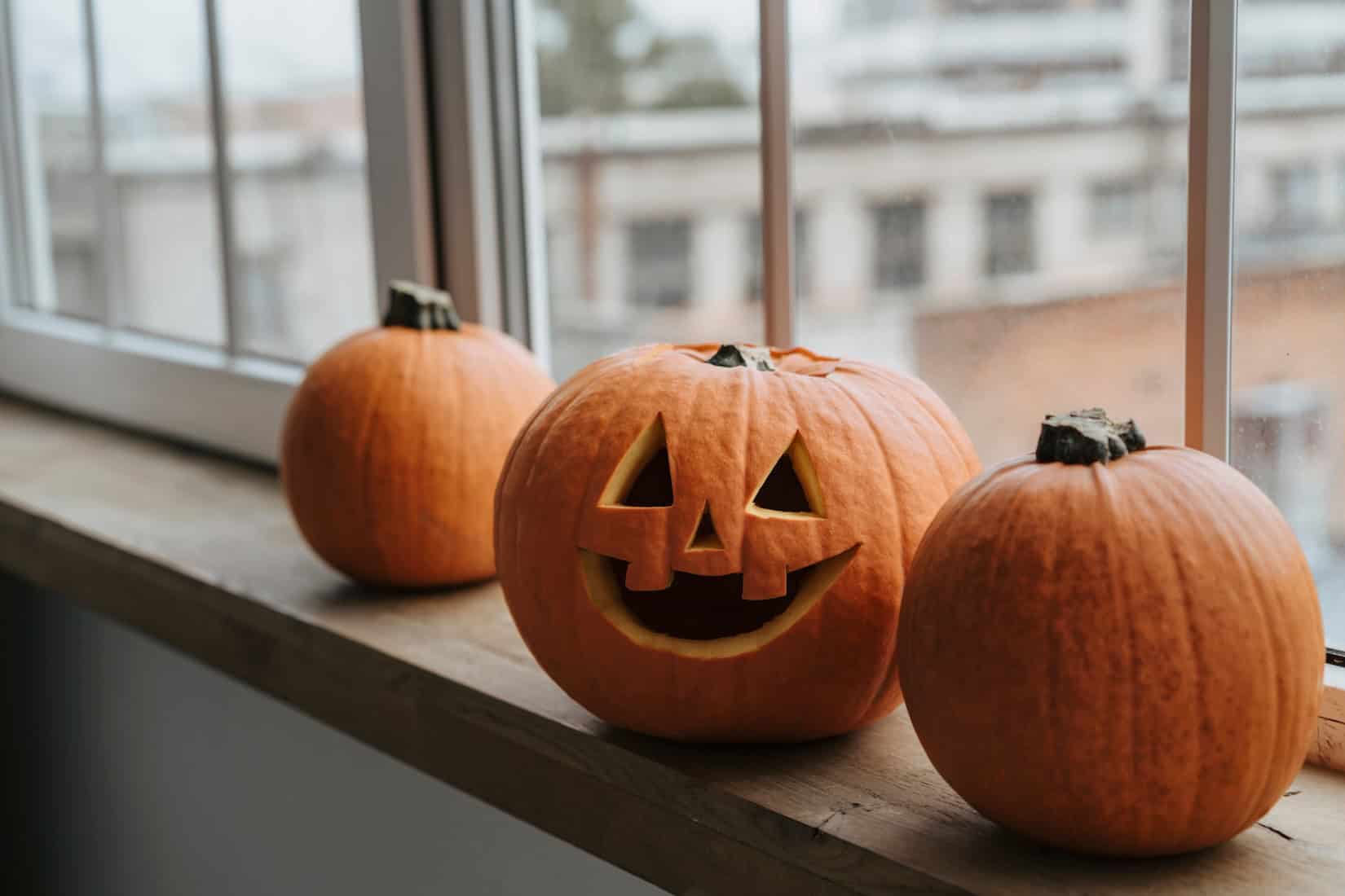 Three pumpkins on a windowsill, one carved with a jack-o'-lantern face.