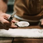 Person holding a fresh oyster with a fork, ready to eat, on a wooden table.