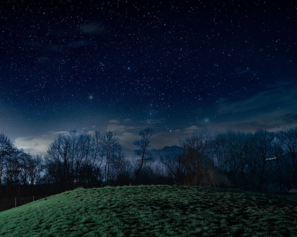 Clear night sky filled with stars over a landscape with trees and hills.